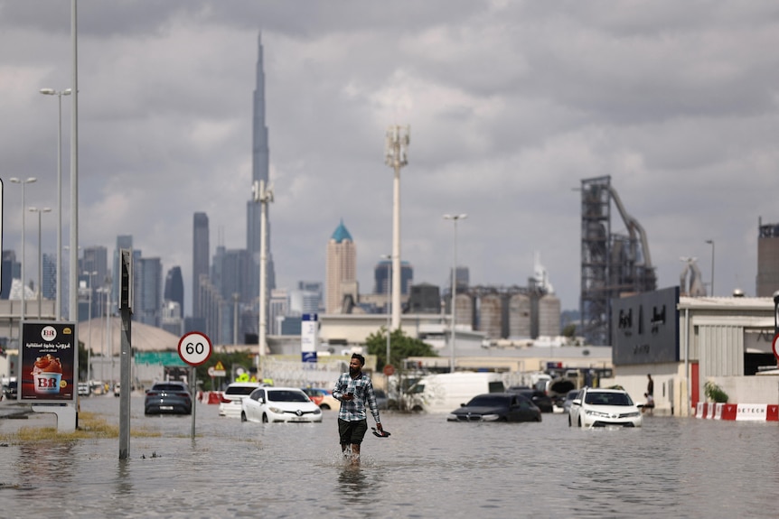 A man walks through flood waters on a road, with cars submerged and a tall tower in the distance
