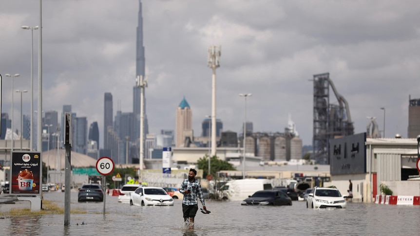 A man walks through flood waters on a road, with cars submerged and a tall tower in the distance