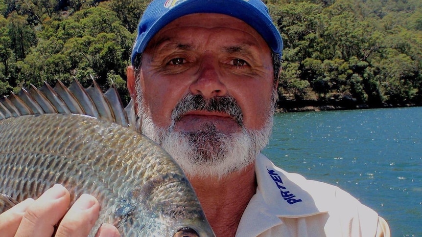 An older, bearded man in a cap holds up a fish while standing in front of a waterway.
