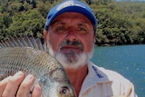 An older man with a salt-and-pepper beard holds up a fish while standing in front of a waterway.