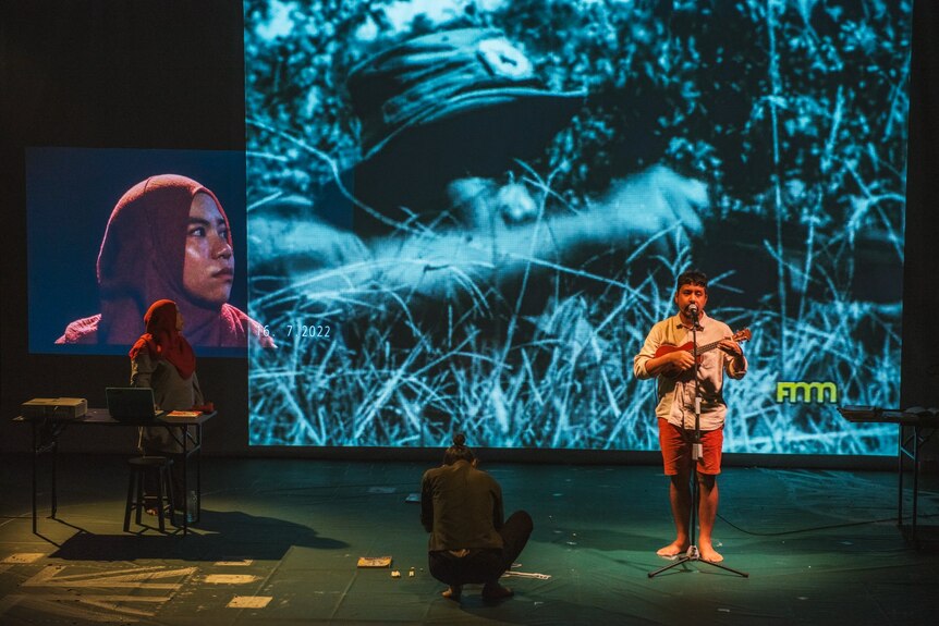 Three Malaysian performers are onstage: a woman in a hijab, a man crouching drawing on the floor, and a man playing the ukelele.