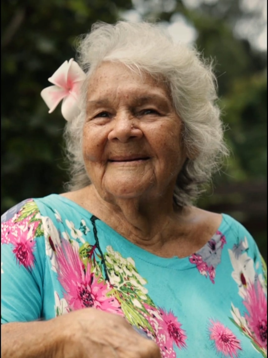 A portrait of Aboriginal elder Kathy Mills, smiling, with a pink frangipani in her hair outside on a sunny day.