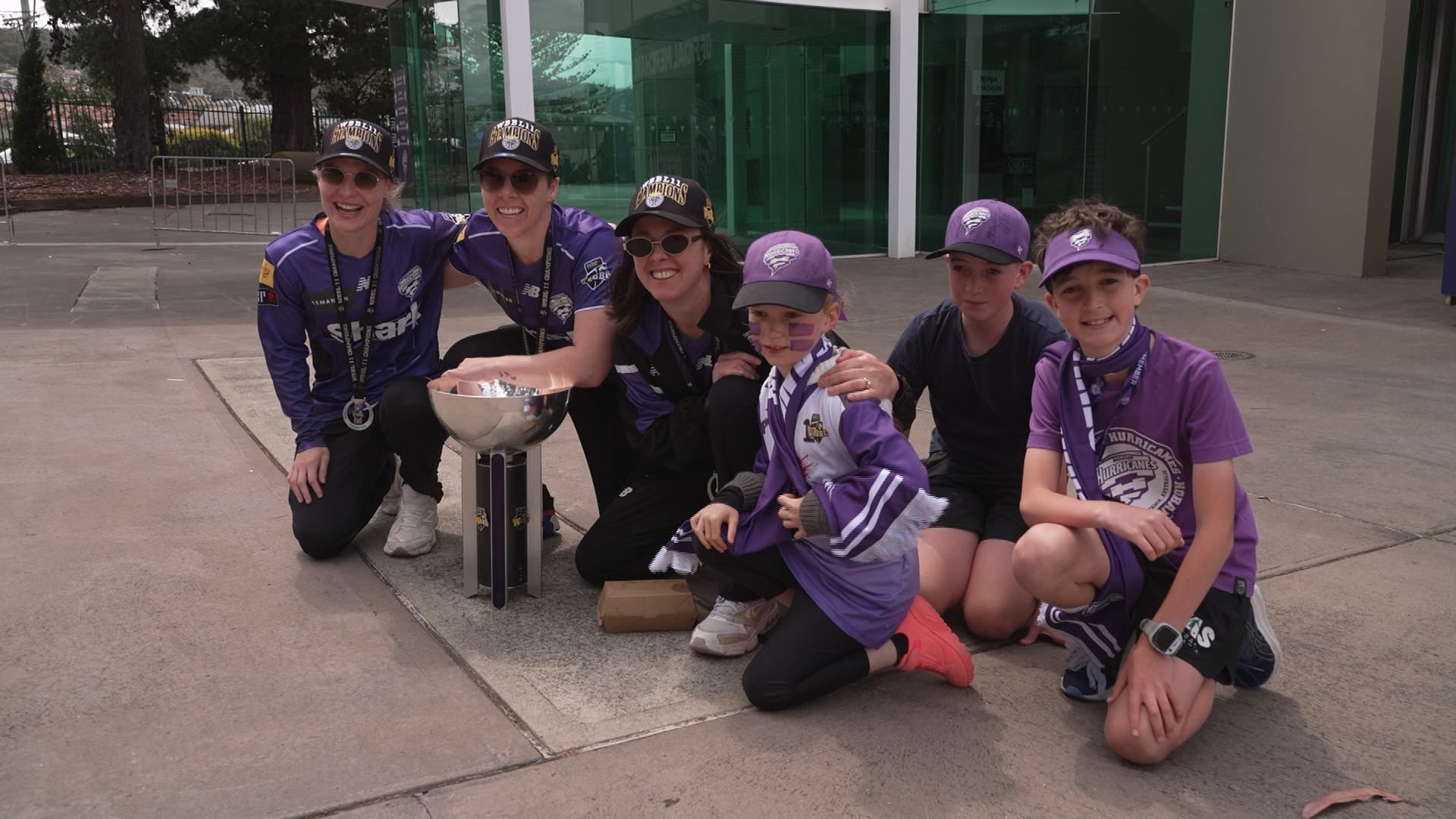 Three Hobart Hurricanes women's players with a trophy and three kids all dressed in Hurricanes' gear kneeling down.