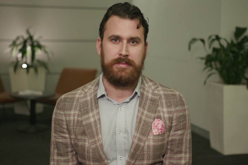 Nick Fischer, a man with a brown beard who is wearing a checked blazer, poses for a photo in an office space