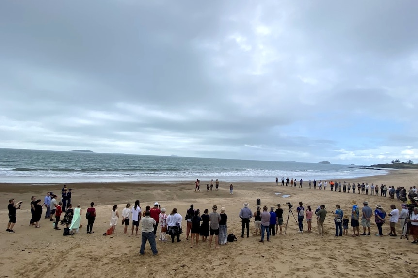 A circle of people in a crowd on a sandy beach. The sky is gloomy