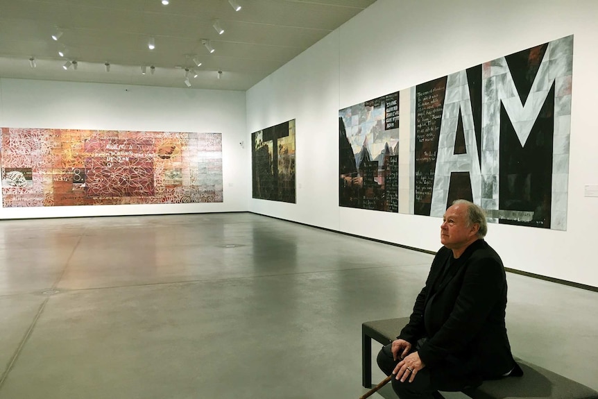 An older man with a walking stick sits in a gallery filled with his paintings