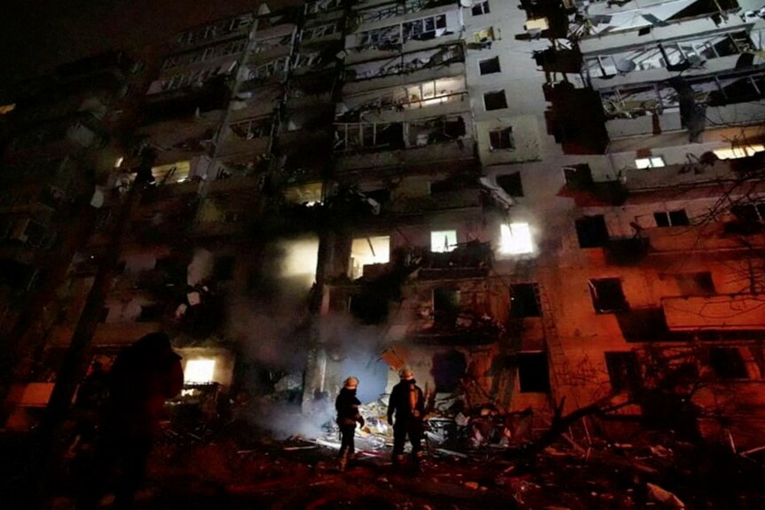Two people stand in front of a large, damaged residential building in Kyiv, Ukraine.