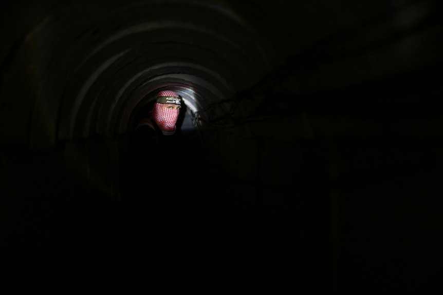The face of a Hamas fighter lit by a torchlight while the rest of the underground tunnel is surrounded by darkness.