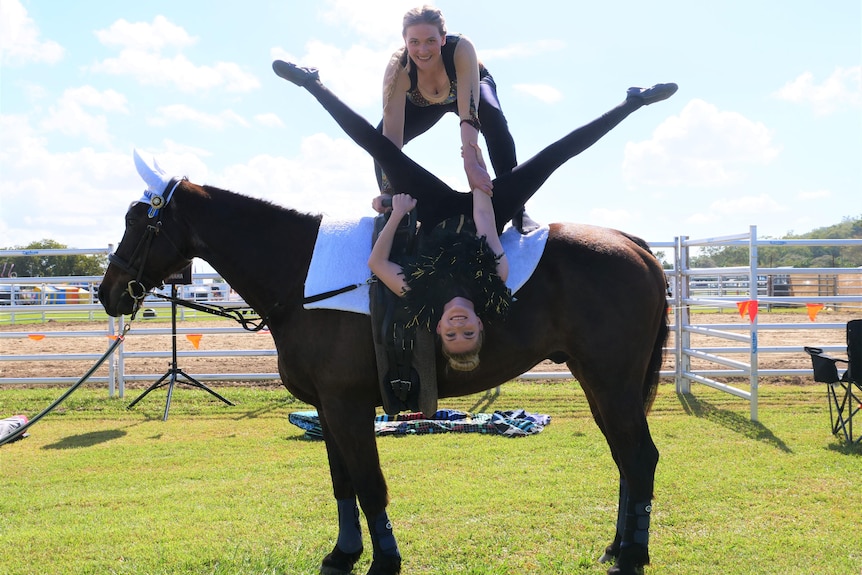 two young women on a horse, one hanging upside with legs in splits while the other stands holding her arm