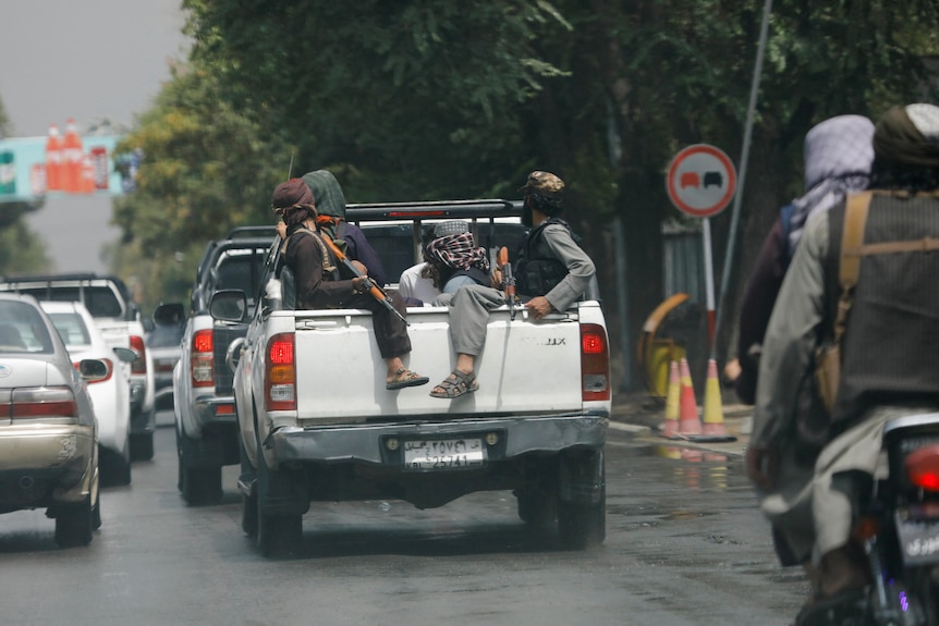 Men holding guns sit in the back of a car on a crowded street.