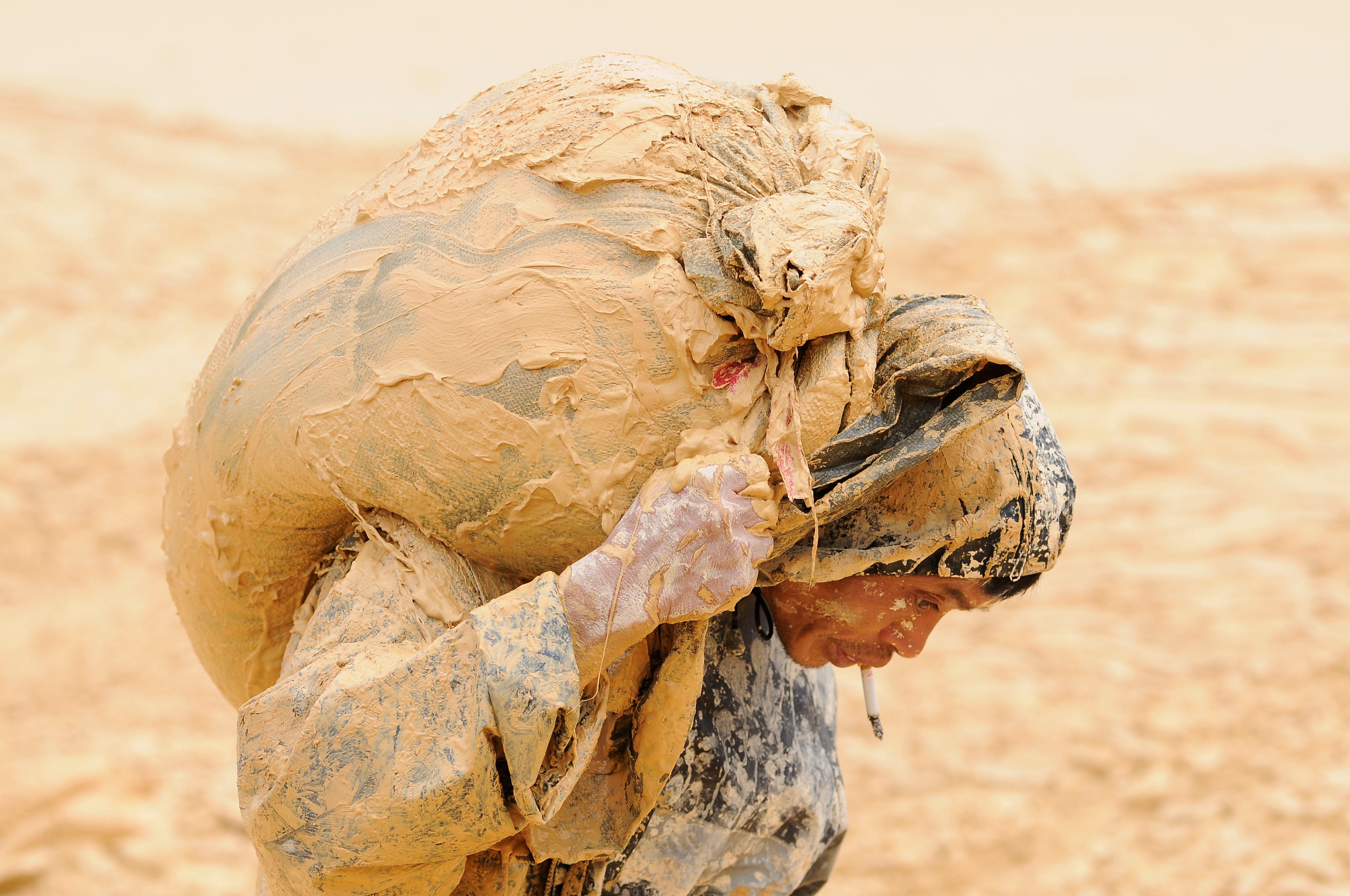 A man covered in mud carrying a sack on his back while smoking a cigarette.