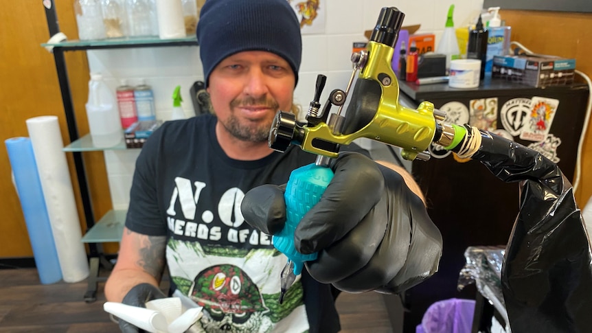 A man sits in a tattoo studio holding a tattoo gun. 