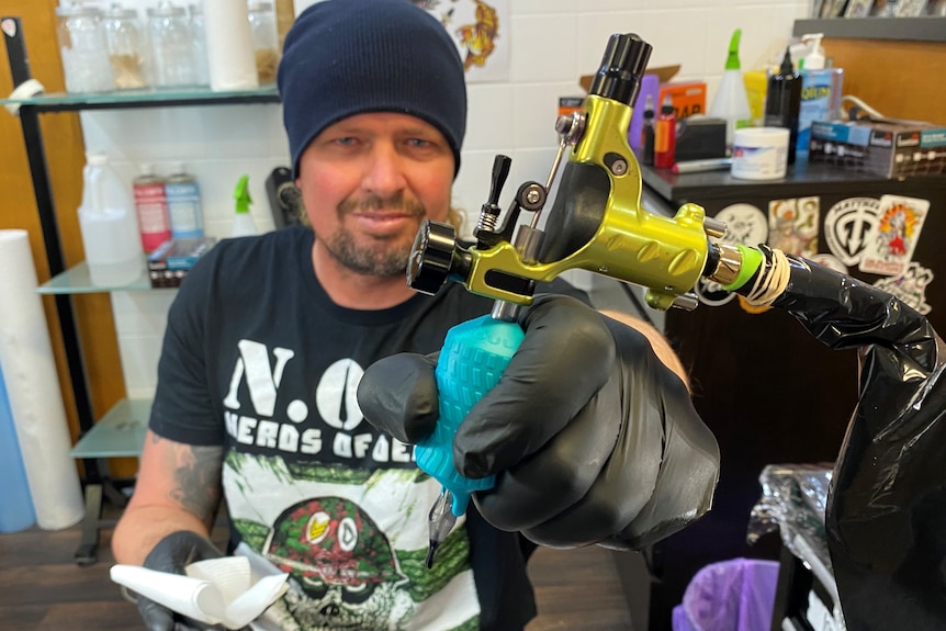 A man sits in a tattoo studio holding a tattoo gun. 