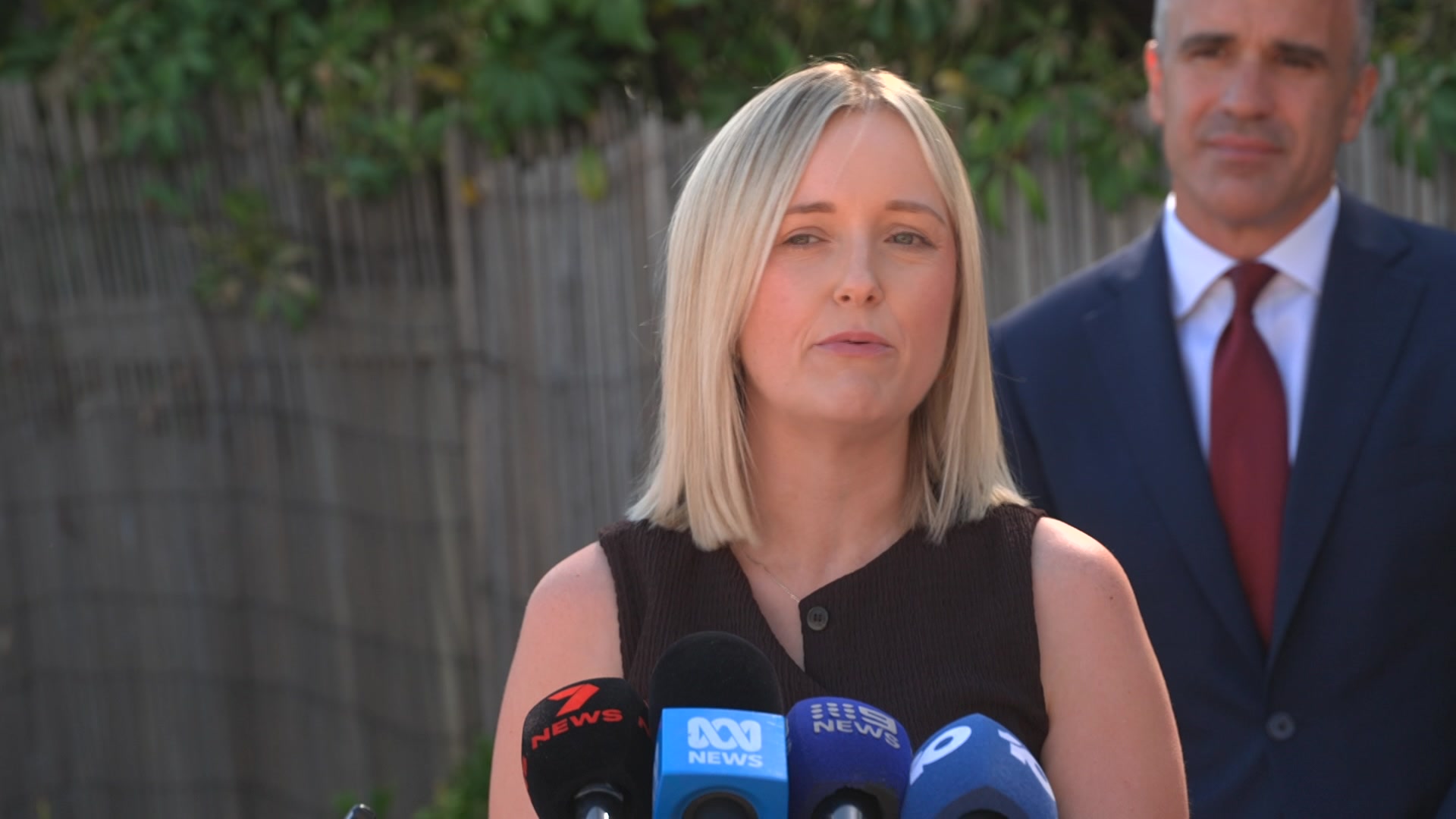 Ellie Simmons tem cabelos loiros na altura dos ombros e quatro microfones de notícias à sua frente enquanto fala.