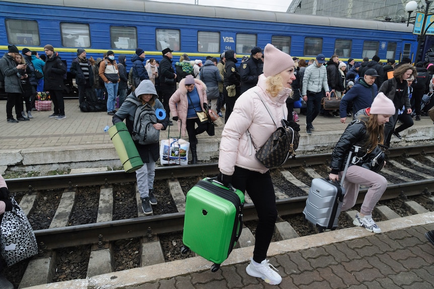 A large crown of people, many with suitcases, line along a train stattion platform while others cross the tracks.