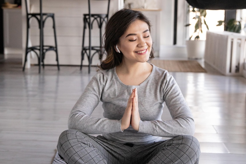 A woman sits cross legged indoors on a mat, palms pressed together, listening to earbuds and smiling. 