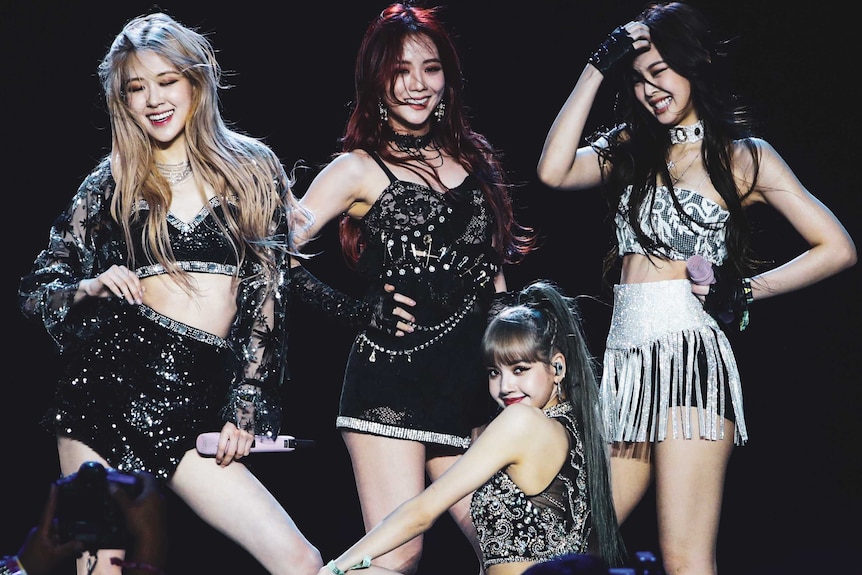 Four women of Asian appearance stand on a stage against a black background.