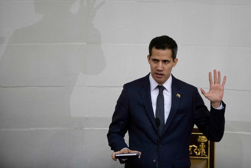 Juan Guaido holds up his left hand as he stands in front of a white wall.