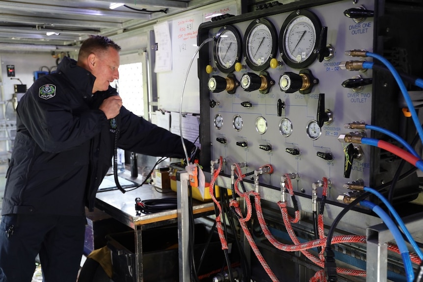 A machine with valves, wires and gauges on the right, a man talking into a walkie-talkie in a police uniform on the left.