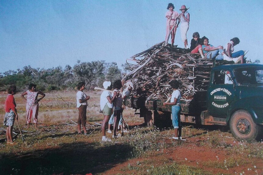Indigenous children stand around a vintage truck piled high with sticks, some children are on top of the load.