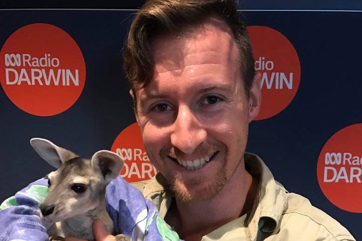 Man smiling holding a baby wallaroo in a blanket with ABC Radio Darwin logo behind.