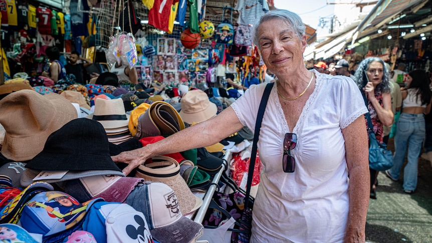 An older woman with cropped grey hair grins while reaching for a hat at a market stall 