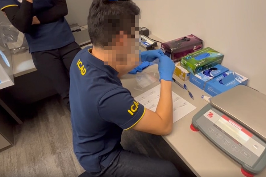 A man with a pixelated face at a desk with blue gloves on 