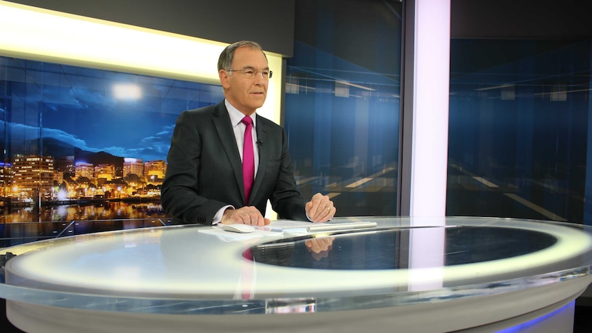 Gee in  suit sitting behind news desk preparing to read the news.