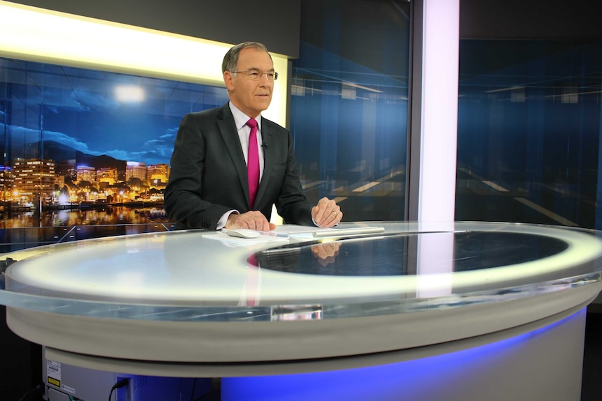 Gee in  suit sitting behind news desk preparing to read the news.