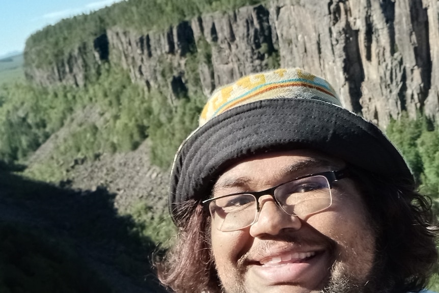 a happy looking man in glasses and a bucket hat smiles in a selfie with a rock landscape in the background.