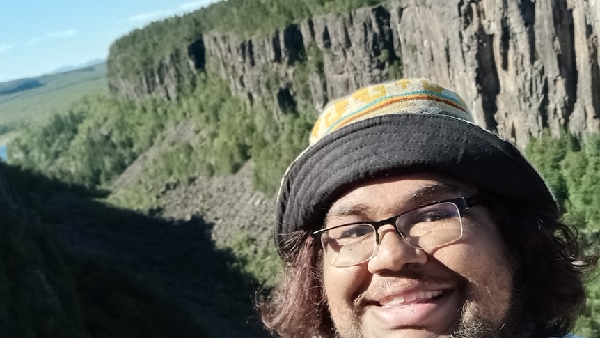 a happy looking man in glasses and a bucket hat smiles in a selfie with a rock landscape in the background.
