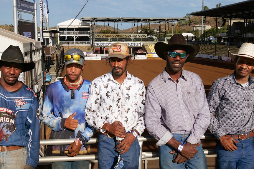 A group of Aboriginal rodeo riders wearing cowboy gear pose in front of a large, dusty arena