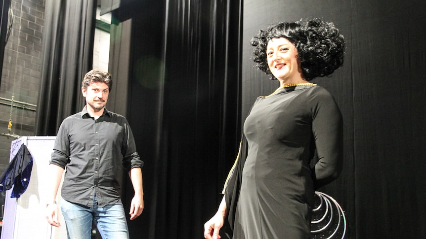 A man in jeans and a woman in a black dress and wig stand on a theatre stage.