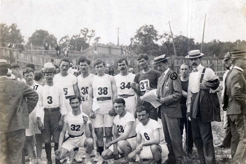A black and white photo of men dressed in running gear surrounded by men in suits