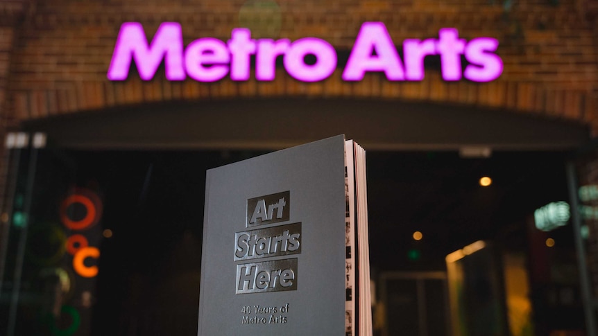 A grey book titled "Art Starts Here" standing on a table, with the entrance to Metro Arts behind it.