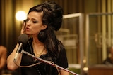Amy holds a headphone ear to her ear as she sings into a microphone, wearing signature eyeliner and beehive.