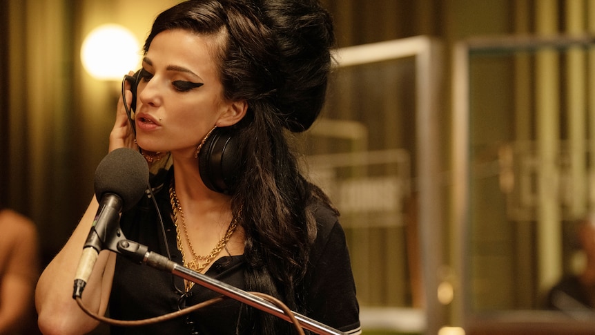 Amy holds a headphone ear to her ear as she sings into a microphone, wearing signature eyeliner and beehive.