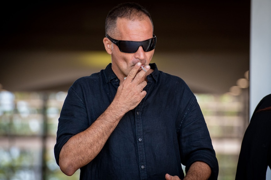 A white man wearing sunglasses smoking a cigarette outside court