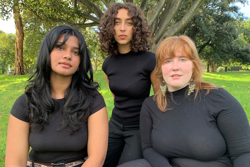 Sinead, Antoinette and Mia are dressed in black and sit in a park on a sunny Melbourne day.
