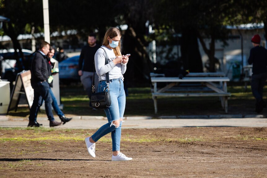 A woman wearing a face mask walks inside Claremont Showground looking at her mobile phone.