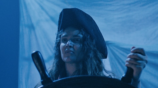 A woman with long dark curly hair wears black pirate holds steering wheel of colonial boat on a dark blue-hued night.