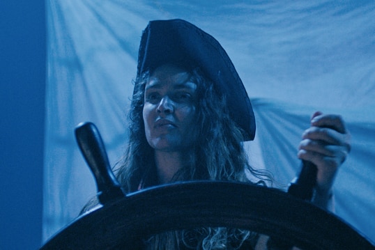A woman with long dark curly hair wears black pirate holds steering wheel of colonial boat on a dark blue-hued night.