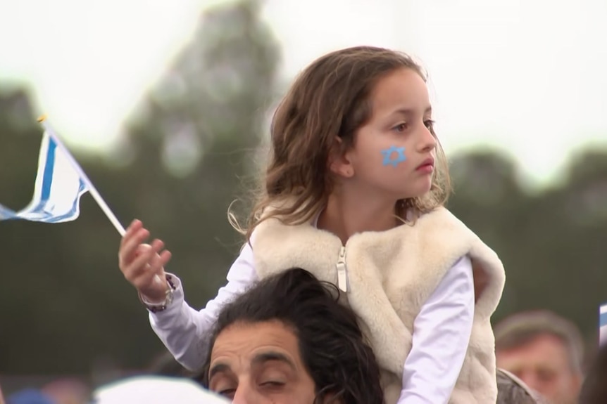 Girl with Star of David painted on her cheek sits on a man's shoulders waving Israeli flag