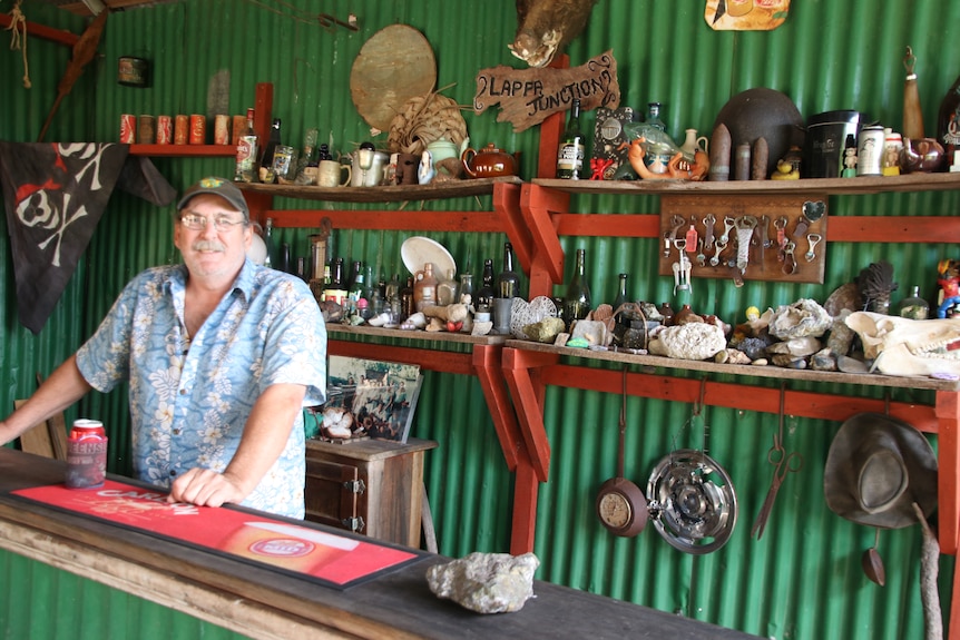 A man stands behind an old wooden bar with ramshackle shelves lined with empty bottles and rocks