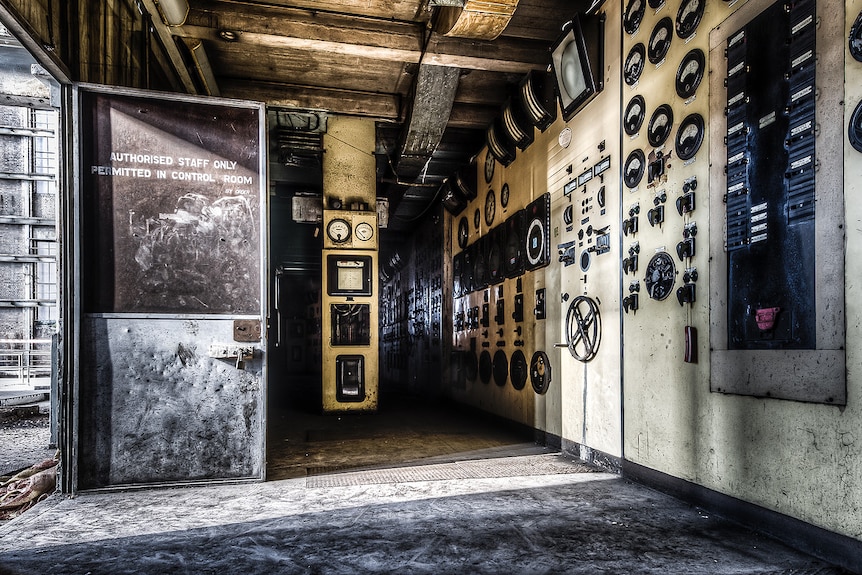 A dark photograph of a boilerhouse control room with a heavy steel door and panels with lots of knobs, dials and wheels