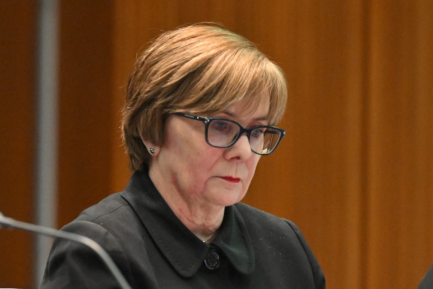 A close up of a middle-aged white woman with short hair, glasses and lipstick on, looking sternly downward