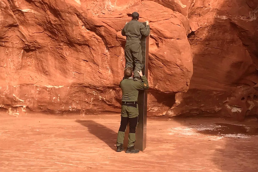 A man stands on another man's shoulders to demonstrate the height of a silver object in a red rock canyon