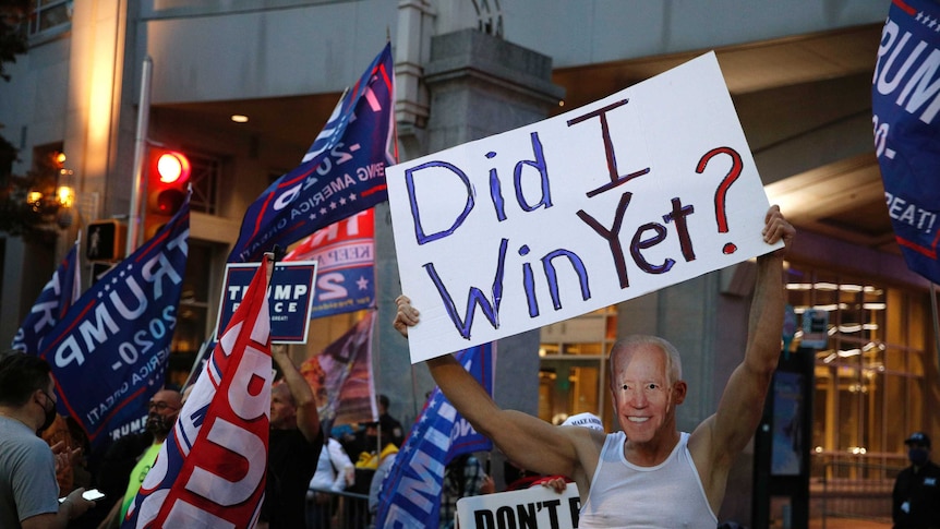 A Trump supporter wears a mask made from a photograph of Joe Biden holding a sign reading 'did I win yet?'