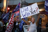 A man wearing a singlet holds up a sign that says 'Did I win yet?'