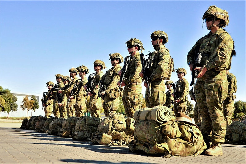 Soldiers in uniform stand in a straight line. They are holding weapons and have their packs in front of them.